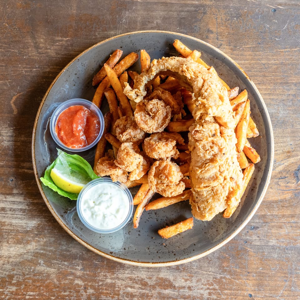 A plate of golden-brown fried fish, shrimp, and french fries, with tartar sauce and ketchup.