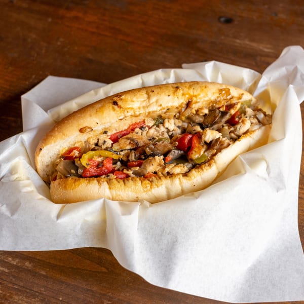 Chicken cheesesteak sandwich with peppers, onions, and mushrooms in a white paper liner on a wooden table.