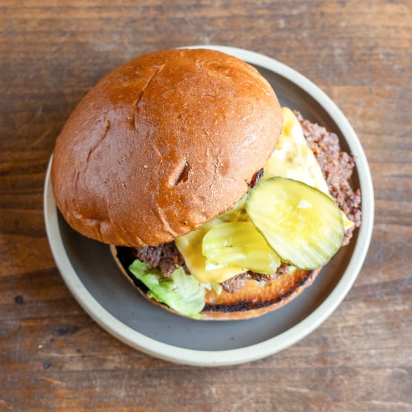 Top-down view of a cheeseburger with pickles and lettuce on a dark plate, on a wooden table.