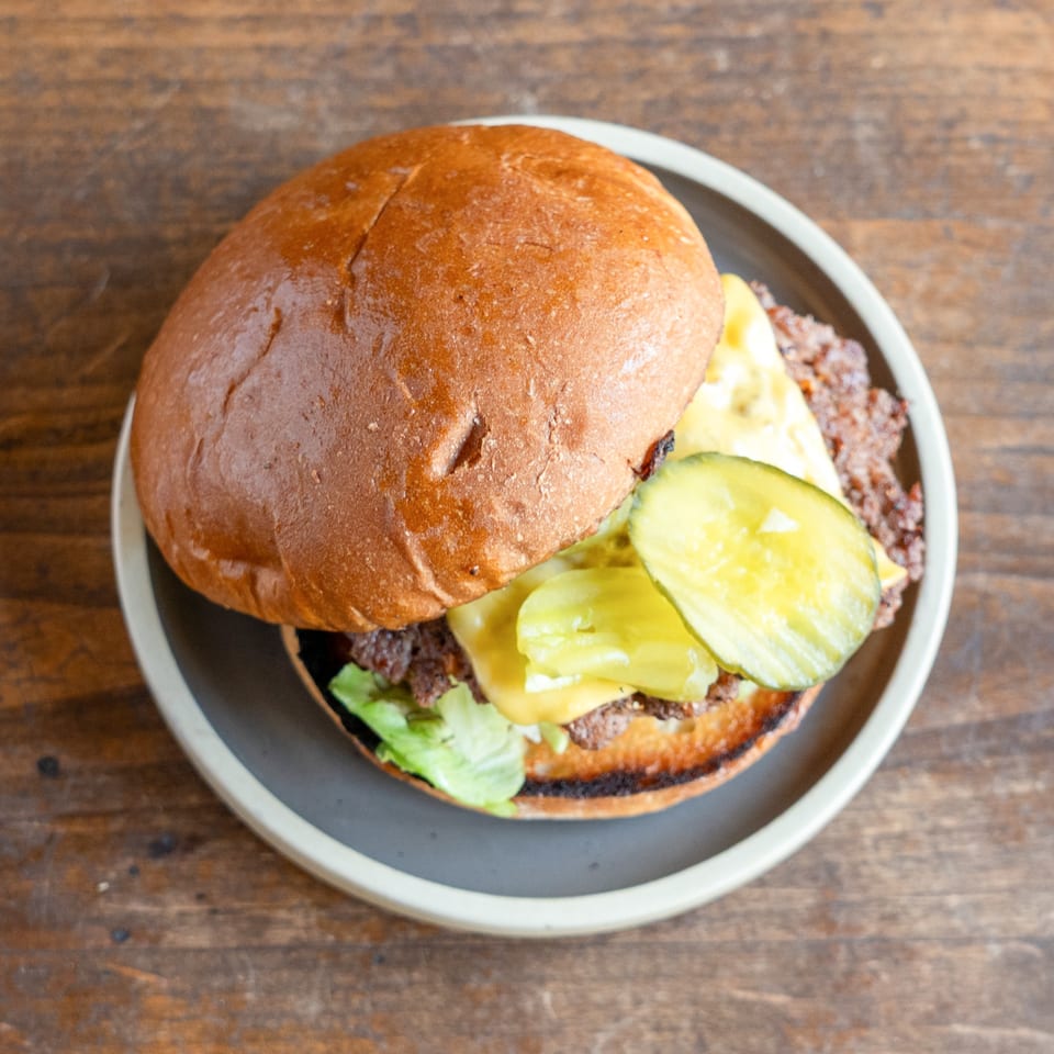 Top-down view of a cheeseburger with pickles and lettuce on a dark plate, on a wooden table.
