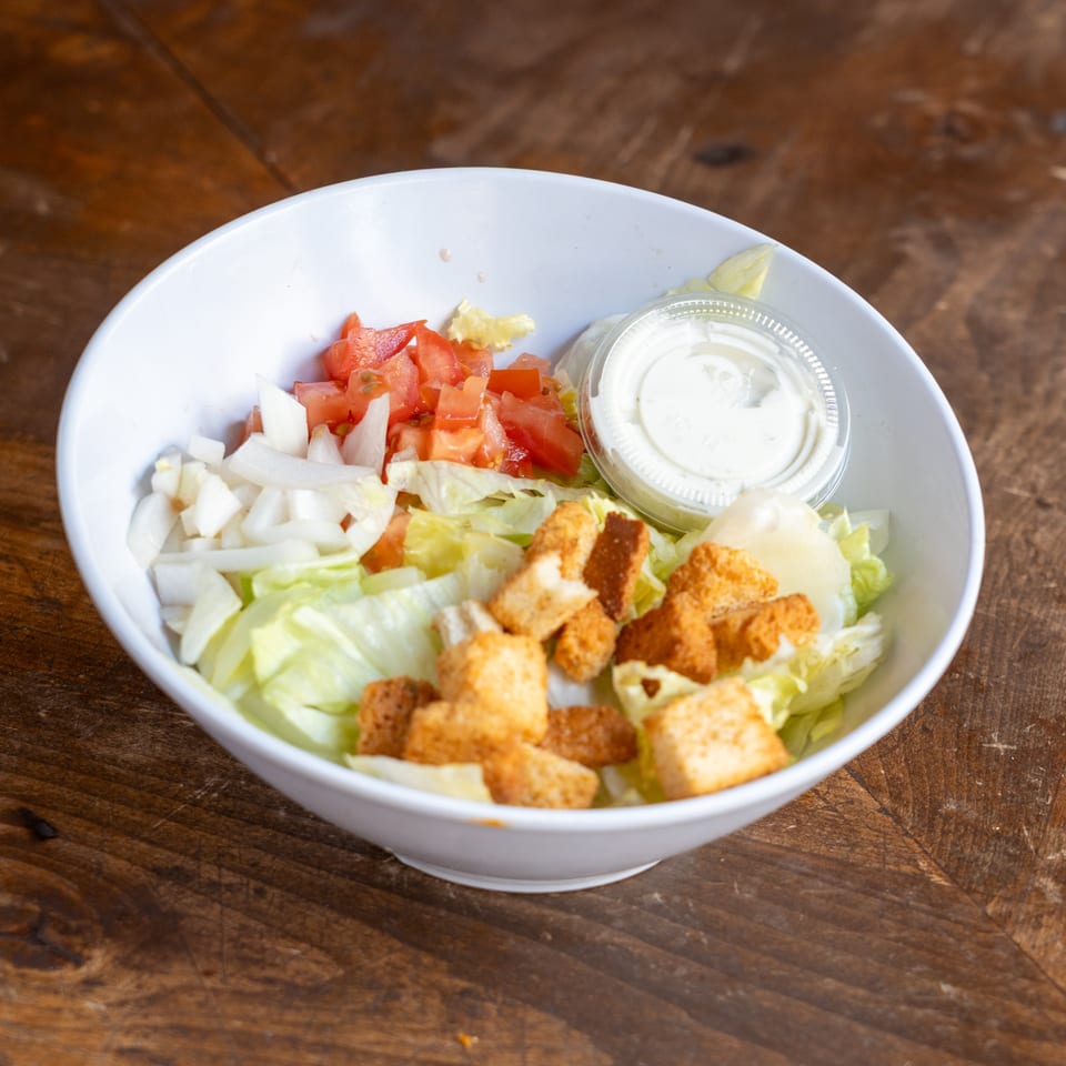 A white bowl of salad with lettuce, diced tomatoes, onions, croutons, and a side of dressing.