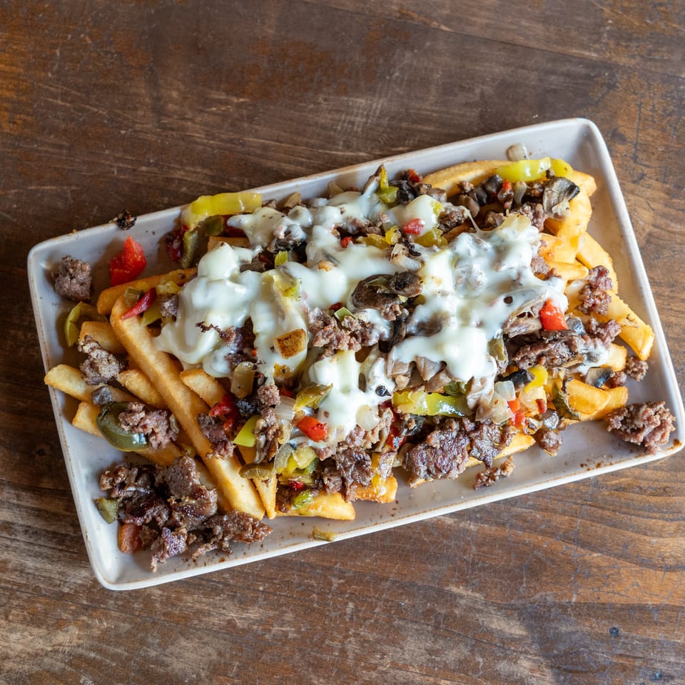 Cheesesteak fries topped with melted cheese, steak, peppers, onions, and mushrooms on a rectangular plate.