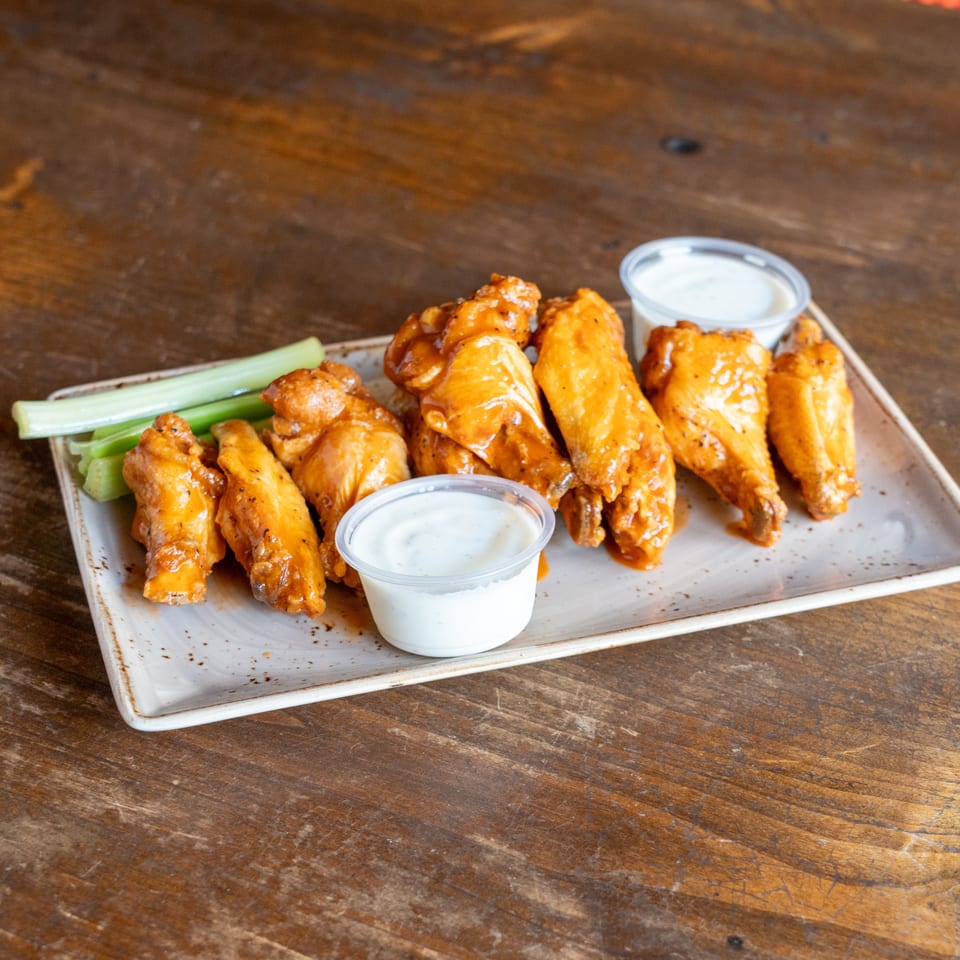 A plate of buffalo chicken wings with celery sticks and two cups of ranch dressing on a wooden table.