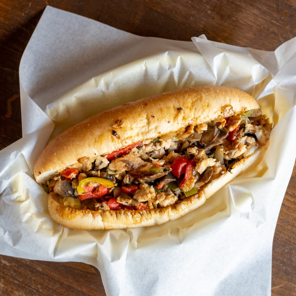 A chicken cheesesteak sandwich with mushrooms, red and green peppers, and onions in a paper-lined basket.